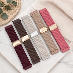May include: Five braided elastic watch bands in burgundy, dark brown, light gray, beige, and pink. Each band features a rectangular metal slider buckle with a smooth finish. The bands are arranged side-by-side, showcasing the woven texture.