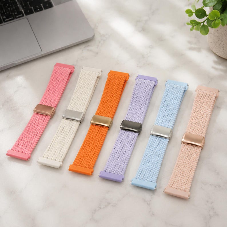 May include: Six braided watch bands in pink, white, orange, lavender, light blue, and beige. Each band features a rectangular metal sliding buckle for adjustment. The bands are arranged in a row, showcasing a woven texture and metallic hardware.