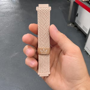 May include: A light pink braided watch band featuring a rectangular metallic rose gold slider with a white line-art crown icon engraved on the front. The band has matching light pink plastic connectors at both the top and bottom ends.