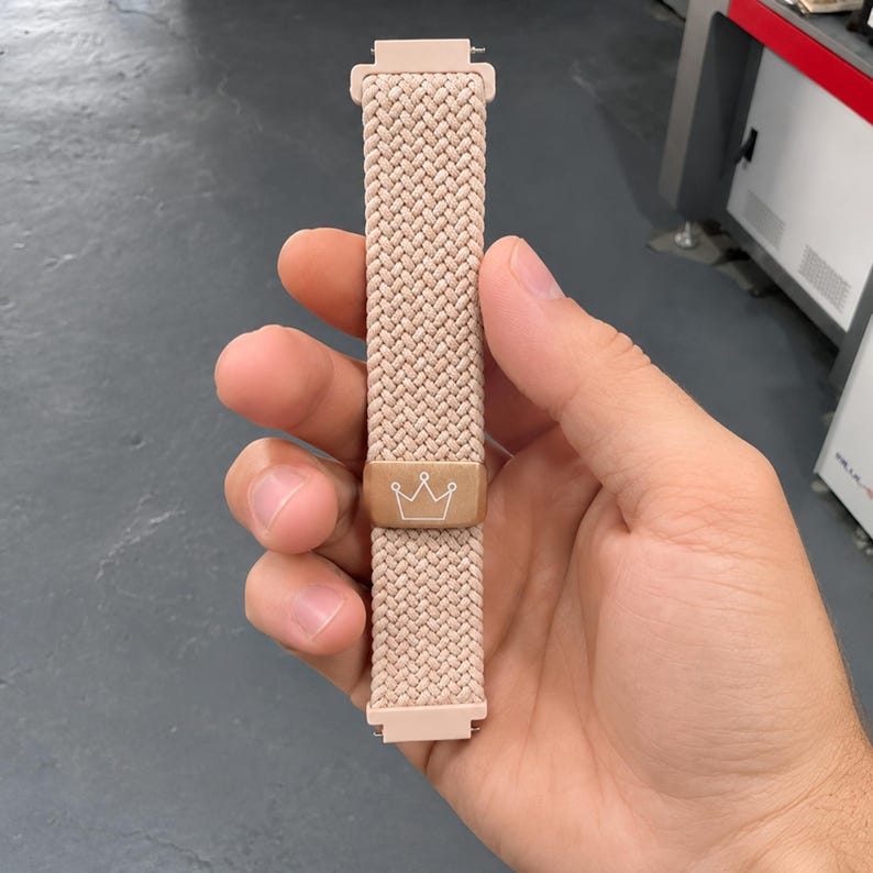May include: A light pink braided watch band featuring a rectangular metallic rose gold slider with a white line-art crown icon engraved on the front. The band has matching light pink plastic connectors at both the top and bottom ends.