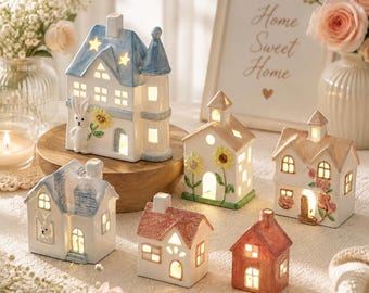 May include: A collection of six ceramic house-shaped tea light holders in various sizes and colors. The houses feature cut-out windows, painted floral and rabbit motifs, and soft interior lighting. Some have blue, pink, or brown roofs.