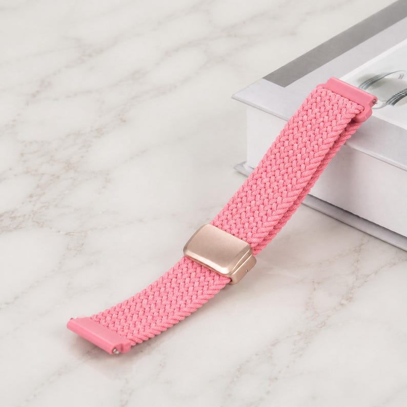 May include: A pink braided watch band featuring a woven fabric texture and a metallic rose gold-toned sliding adjustment buckle. The strap includes integrated connectors at both ends for attachment to a watch face, showcasing a flexible design.