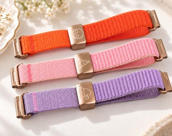 May include: Three fabric watch bands in orange, pink, and purple. Each band features a metallic buckle and a sliding metal loop engraved with a specific icon: a skull on the orange band, a Christmas tree on the pink, and a hat on the purple.