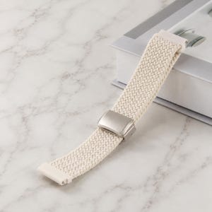 May include: A cream-colored woven watch band featuring a braided texture and a rectangular silver-toned metal sliding buckle. The strap is constructed from a flexible material and includes a solid white connector piece at the end for attachment.