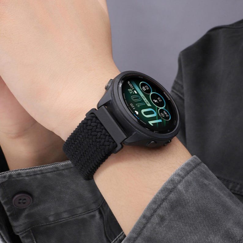 May include: A black smartwatch with a round digital display showing the time 10:10, battery level, and activity metrics. The watch features a textured, braided black elastic strap and a dark casing with side buttons, worn on the wrist.