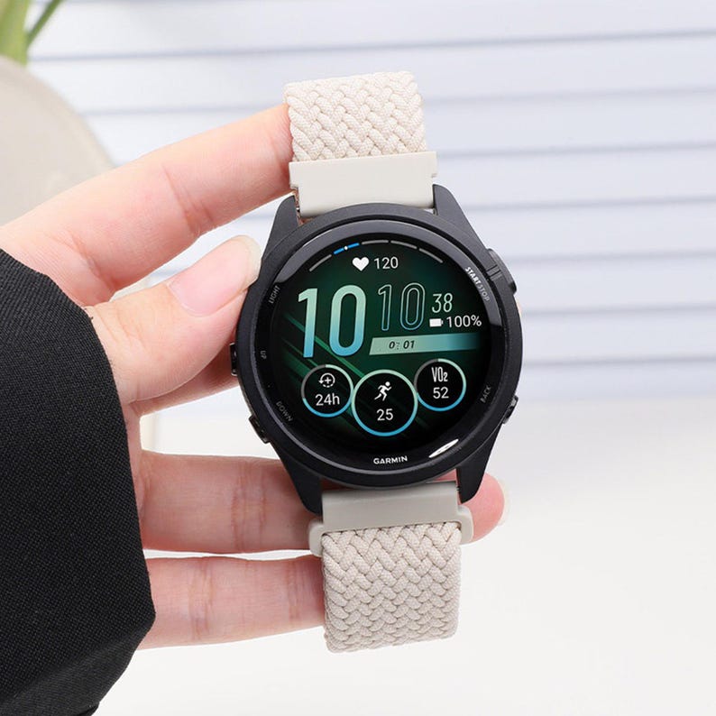 May include: A black Garmin smartwatch with a round digital display showing the time 10:10, a heart rate of 120, a 100% battery level, and other fitness metrics. The watch features a textured, beige woven fabric strap with light-colored plastic connectors.