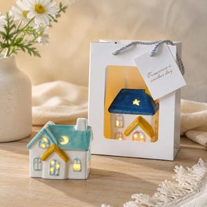 May include: Two ceramic house-shaped tea light holders. One is teal and white with a yellow roof accent and moon cutout, sitting outside a white gift bag containing a second house with a blue roof, star cutout, and the text Tomorrow is another day.
