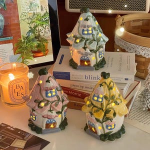 May include: Three ceramic house-shaped tea light holders in pastel blue, pink, and yellow. Each structure features a pointed roof, small arched windows, and green vine details. The houses are illuminated from within, casting a warm glow.