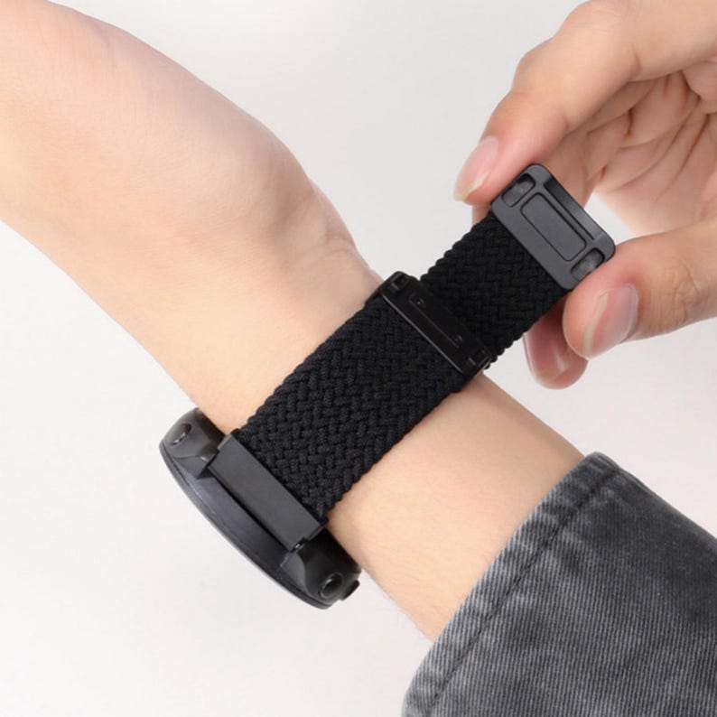 May include: A black braided watch band featuring a textured woven pattern and a solid black rectangular buckle mechanism. The strap is attached to a circular black watch casing, showcasing a flexible design that wraps around the wrist area.