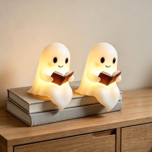 May include: Two white, glowing ghost-shaped lamps, each featuring a smooth, rounded head, black oval eyes, and a small smile. Each ghost figure is holding a small, open brown book in its hands, emitting a soft, warm light from within its body.