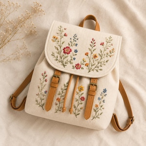 May include: A cream-colored canvas backpack featuring intricate floral embroidery in shades of red, yellow, and blue. The bag includes tan faux leather straps with metal buckles, a top handle, and a drawstring closure, accented by delicate green stems.