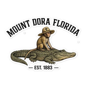May include: A sticker featuring a brown monkey wearing a straw hat while riding on the back of a green alligator. The text MOUNT DORA FLORIDA is arched above the monkey, and the text EST. 1883 is centered below the alligator in black font.