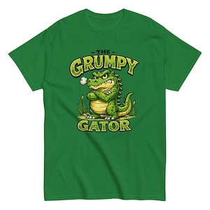 May include: A bright green short-sleeve t-shirt featuring a graphic of a grumpy cartoon alligator with crossed arms and a puff of steam from its nose. The text THE GRUMPY GATOR is printed in bold, stylized green letters above and below the character.