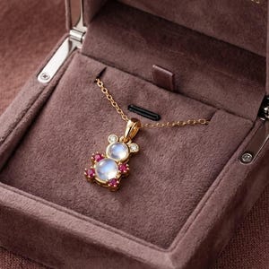 May include: A gold necklace featuring a bear-shaped pendant. The pendant is set with two round moonstones for the head and body, two small clear diamonds for the ears, and four small red rubies accenting the limbs, all in a gold setting.