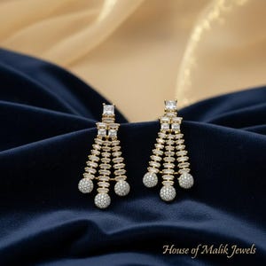 May include: A pair of gold-toned chandelier earrings featuring a fan-like arrangement of baguette-cut clear stones, accented with square-cut stones at the top and three round, pavé-set stone spheres at the base. The text House of Malik Jewels is visible.