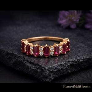 Puede incluir: Un anillo de oro con un patrón alterno de rubíes rojos rectangulares en talla baguette y pequeños diamantes blancos redondos en talla brillante. Las piedras están engastadas en una fila continua en la parte superior de la banda de oro.