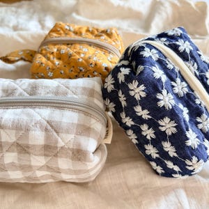 May include: Three small, quilted zipped pouches. One is mustard yellow with a white floral print, one is beige and white gingham, and one is navy blue with a white embroidered daisy pattern. Each pouch features a zip closure and a small fabric tab.