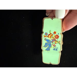 May include: A rectangular mint green brooch with rounded edges and a gold-toned metal border. The surface features a painted design of a blue bird in flight alongside a colorful floral arrangement with red, orange, and blue petals and green leaves.