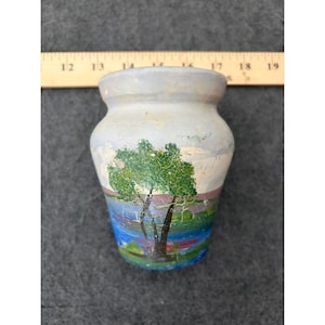 May include: A small, light-colored ceramic vase featuring a hand-painted landscape scene with green trees, a blue body of water, and a purple horizon line. A wooden ruler is positioned behind the vase, showing a width of approximately 2.5 inches.