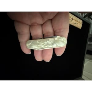 Pode incluir: Um canivete fechado com um cabo branco perolado liso com uma textura marmorizada. O canivete está posicionado ao lado de uma régua de madeira que mostra uma medida de aproximadamente 7,6 cm de comprimento para a seção do cabo.