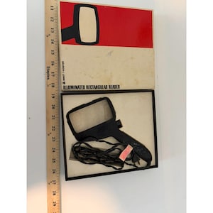 Pode incluir: Um leitor retangular iluminado Bausch & Lomb com cabo e moldura pretos, incluindo um cabo de alimentação preto. O dispositivo está alojado numa caixa branca com detalhes vermelhos e o texto "ILLUMINATED RECTANGULAR READER". Uma régua de madeira mostra que a caixa tem cerca de 28 cm de comprimento.