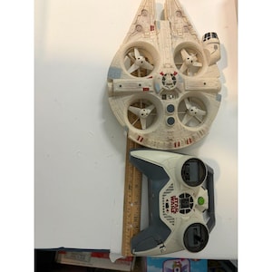 Puede incluir: Un dron teledirigido Millennium Falcon con cuatro hélices blancas y un mando a distancia gris y blanco con el logotipo de Star Wars. Una regla de madera está colocada entre los objetos, mostrando una longitud de unos 28 cm.