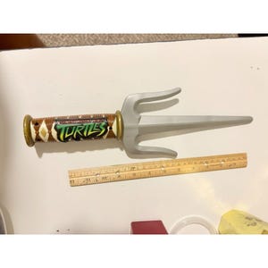 May include: A silver-colored plastic sai weapon featuring a brown handle with a diamond pattern and the Teenage Mutant Ninja Turtles logo. A wooden ruler is positioned below the toy, showing a length of approximately 12 inches for the sai.