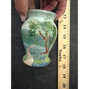 May include: A small, light-colored vase featuring a hand-painted landscape scene with a green tree, a winding path, and blue foliage. A wooden ruler placed alongside the vase indicates a height of approximately 5 inches from base to rim.