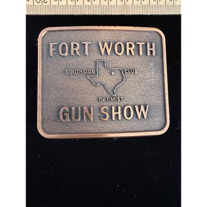 以下が含まれることがあります： 銅色の金属製長方形ベルトバックル。FORT WORTH GUN SHOWの浮き彫り文字、テキサス州の地図、SOUTHSIDE OPTIMIST CLUBの文字が刻まれています。上部の定規により、バックルの幅が約8.9cmであることが確認できます。