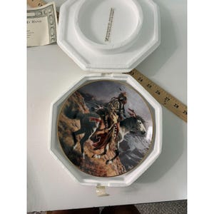 May include: A round decorative plate featuring a detailed illustration of a person riding a horse through a mountainous landscape. The plate is housed in a white styrofoam container with a wooden ruler measuring approximately 23 to 29 inches.
