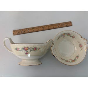May include: A cream-coloured porcelain gravy boat and matching bowl featuring a delicate floral pattern of pink and yellow roses with green leaves and gold trim. A wooden ruler is positioned above the pieces, showing a length of 30 cm.
