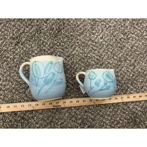 May include: A light blue ceramic pitcher and a matching creamer, both featuring a hand-painted botanical leaf pattern in a darker blue. A wooden ruler is positioned below the items, showing the pitcher is approximately 5.5 inches wide.