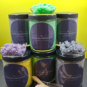 May include: Five clear plastic jars with black lids containing colourful granular scrubs, each featuring a dark purple circular label with the text "COACHED PRODUCTS" and "DEDRIC COTTON". Small clear dishes hold loose scrub samples, and two wooden spoons rest in front.