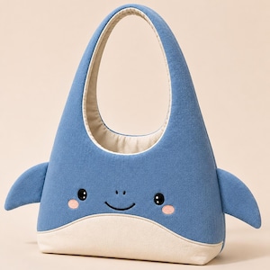 Op de afbeelding: Een blauwe stoffen tas in de vorm van een walvis met een crèmekleurige buik. De tas is voorzien van geborduurde zwarte ogen, een kleine glimlach en twee roze ronde wangen. Twee blauwe zijvinnen steken uit het lichaam en een enkele handgreep.