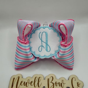 May include: A hair bow featuring pink and light blue horizontal stripes with a pink stitched edge. In the center is a white circular patch with a scalloped edge and a light blue embroidered letter A. Below is a wooden sign reading Newell Bow Co.