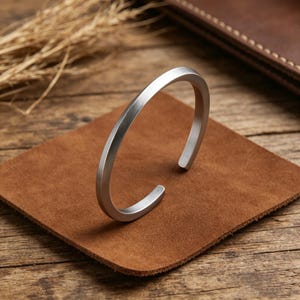 May include: A silver-toned metal cuff bracelet with a smooth, matte finish and a simple open-ended design. The bracelet features a rounded, uniform band that curves into a circular shape, showcasing a clean and unadorned aesthetic throughout.
