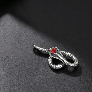 May include: A silver-toned metal snake pendant featuring a coiled body with detailed scale textures and a single round, vibrant red gemstone set into the head of the snake. The design depicts a cobra with its hood flared, showcasing a metallic finish.
