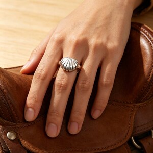 May include: A silver ring featuring a polished, three-dimensional seashell design with vertical ridges. The ring has a smooth, rounded band and a metallic finish that reflects light, showcasing the detailed contours of the shell shape.