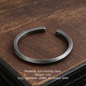 May include: A 925 sterling silver cuff bracelet with a dark, brushed finish. The open-ended circular band has a diameter of 56mm and a thickness of 4mm. The metal surface shows subtle texture and weighs 25g, as indicated by the printed text.