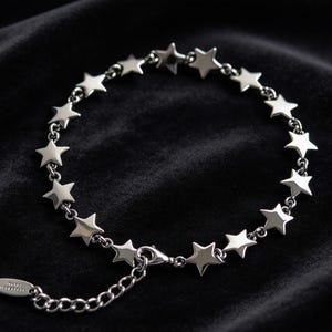 May include: A silver-toned bracelet featuring a series of connected star-shaped charms. The chain includes a lobster claw clasp and an adjustable extender chain with a small, oval-shaped metal tag attached at the end of the links.