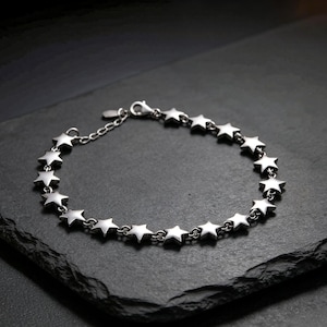 May include: A silver-toned bracelet featuring a continuous chain of small, uniform star-shaped links. The bracelet includes a lobster claw clasp and a short extender chain with a small metal tag attached at the end of the closure mechanism.