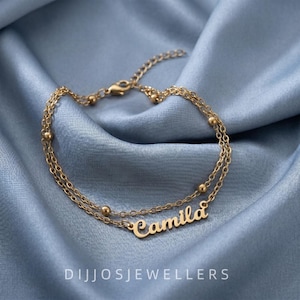 May include: A gold-toned double-chain bracelet featuring small spherical beads along the links and a central nameplate with the name Camila in a cursive script font. The jewelry piece includes a lobster claw clasp and a short extender chain.