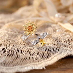 May include: A silver-toned open ring featuring a sunflower motif with orange stones at the center and a bee design with a yellow pear-shaped stone for the body and clear stones on the wings. The text DIJJOSJEWELLERS is visible at the bottom.