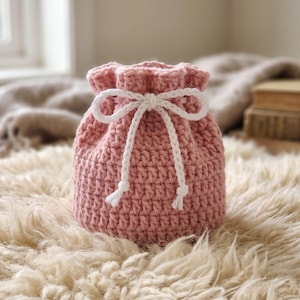 May include: A dusty rose crochet drawstring pouch featuring a textured stitch pattern and a white braided cord closure tied in a bow at the top. The pouch has a rounded base and a gathered opening, providing a compact storage solution for small items.