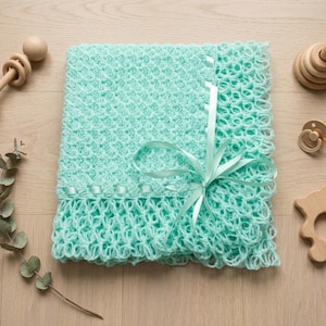 May include: A mint green crocheted baby blanket with a textured diamond pattern and a decorative openwork border. The blanket is tied with a matching mint green satin ribbon bow. Surrounding the blanket are wooden toys, a pacifier, and a eucalyptus sprig.