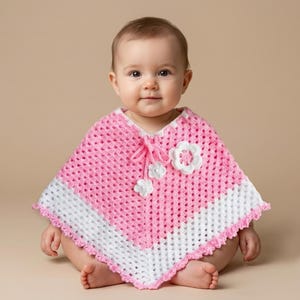 May include: A pink and white crocheted poncho featuring a diamond-patterned knit, a white horizontal stripe near the bottom edge, a decorative drawstring with two small white flower appliqués, and one larger white flower appliqué on the chest.