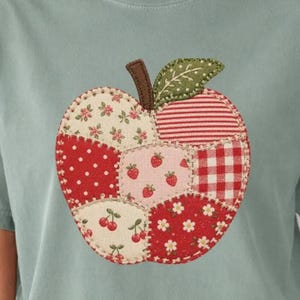 May include: A sage green t-shirt featuring a large, central apple-shaped appliqué. The apple is constructed from various fabric patches in red, white, and cream, displaying patterns of polka dots, gingham, stripes, and floral prints with a brown stem.