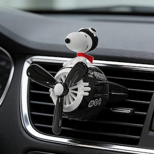 May include: A black and white car air freshener shaped like a vintage aeroplane with a propeller. A Snoopy figure wearing a black aviator cap and a red scarf sits on top. The side of the plane features the text Snoopy and the number #01 in white.