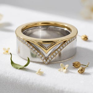 May include: A two-tone ring featuring a wide silver-colored band with a brushed finish and a polished gold-colored top section. The ring is accented with a V-shaped inlay of small, clear, round-cut stones that follow the chevron-style design.