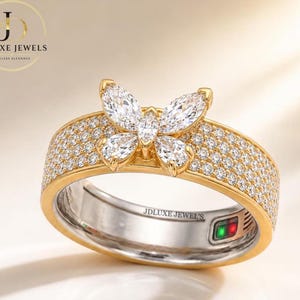 May include: A gold ring featuring a central butterfly motif composed of four marquise-cut clear stones. The band is encrusted with numerous small round clear stones. The interior of the band is engraved with the text JDLUXE JEWELS.
